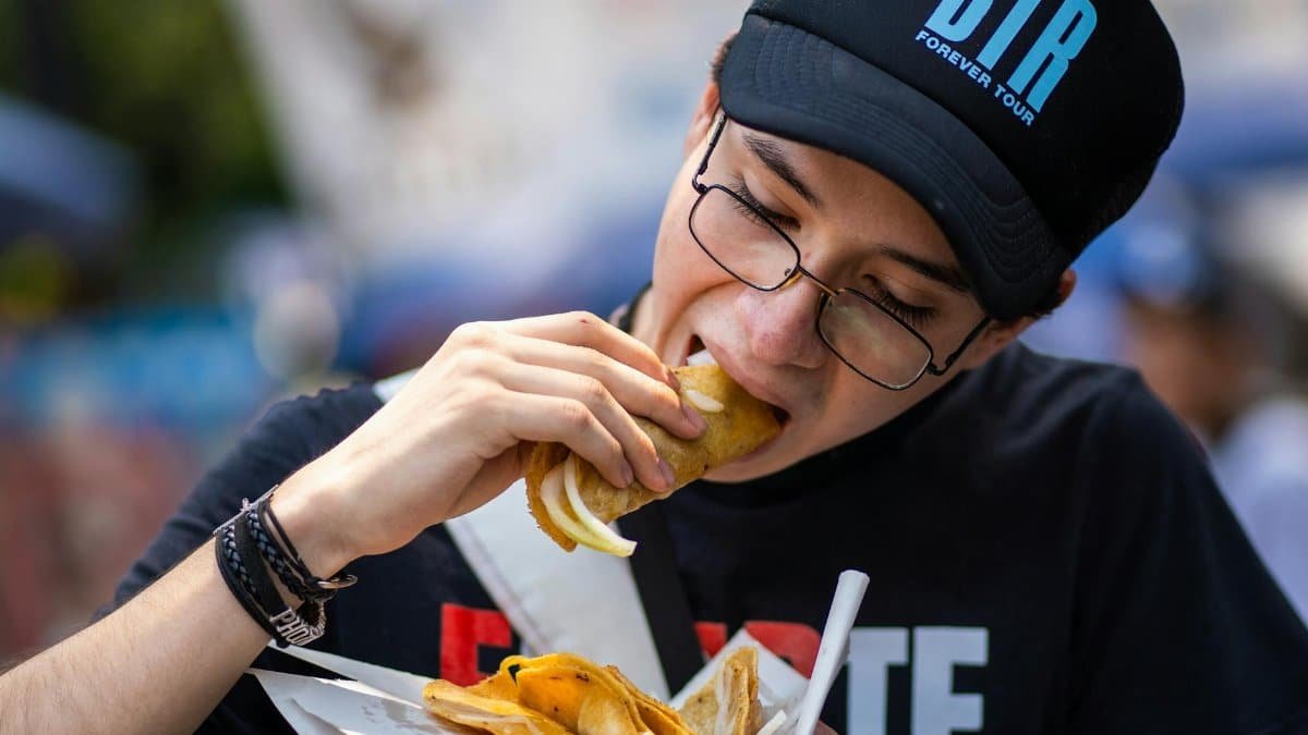 A young man wearing a cap and glasses enjoys street tacos in Mexico City.