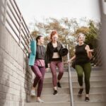 Three women walking down steps outdoors, carrying yoga mats, enjoying a fitness conversation.