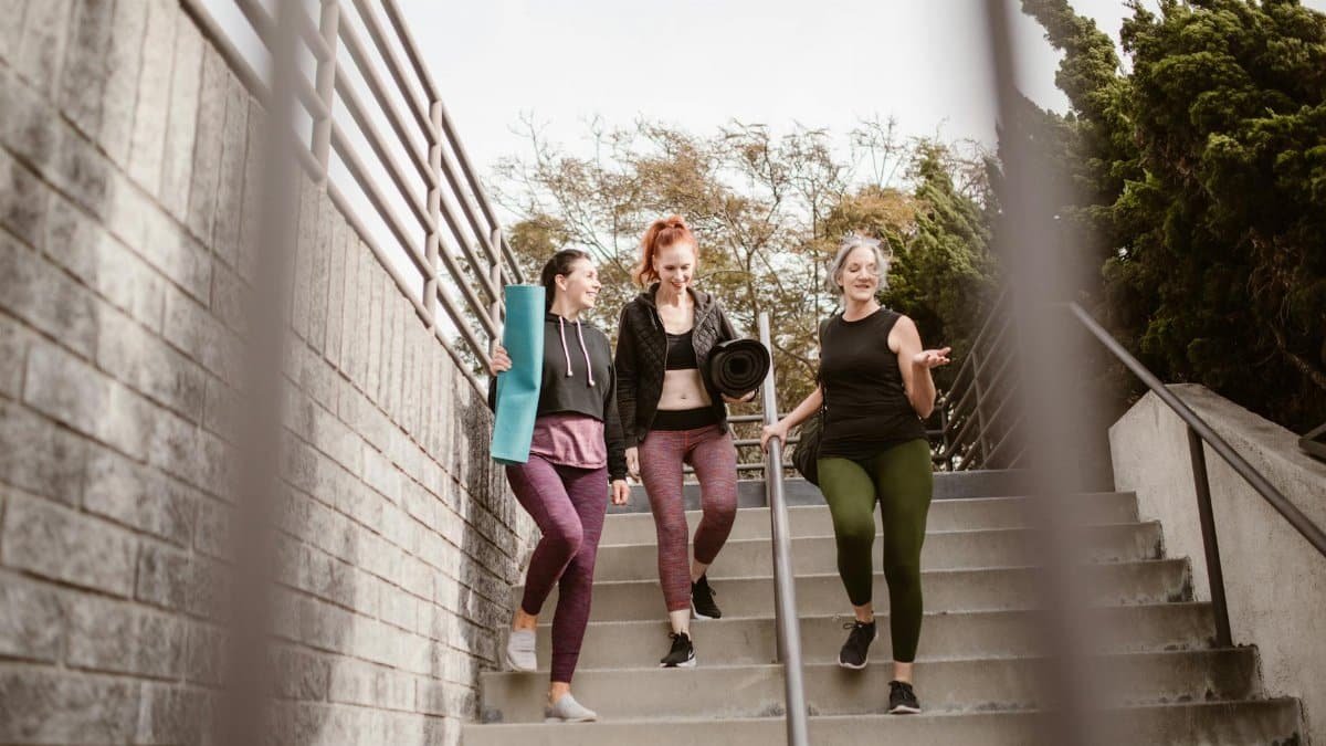 Three women walking down steps outdoors, carrying yoga mats, enjoying a fitness conversation.