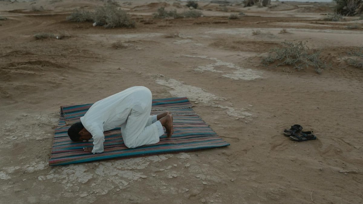 A Muslim man kneels in prayer on a colorful mat in the desert during sunset, showcasing a serene moment of faith.