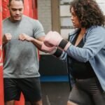 Serious ethnic coach explaining exercise to overweight African American woman punching bag in boxing gym