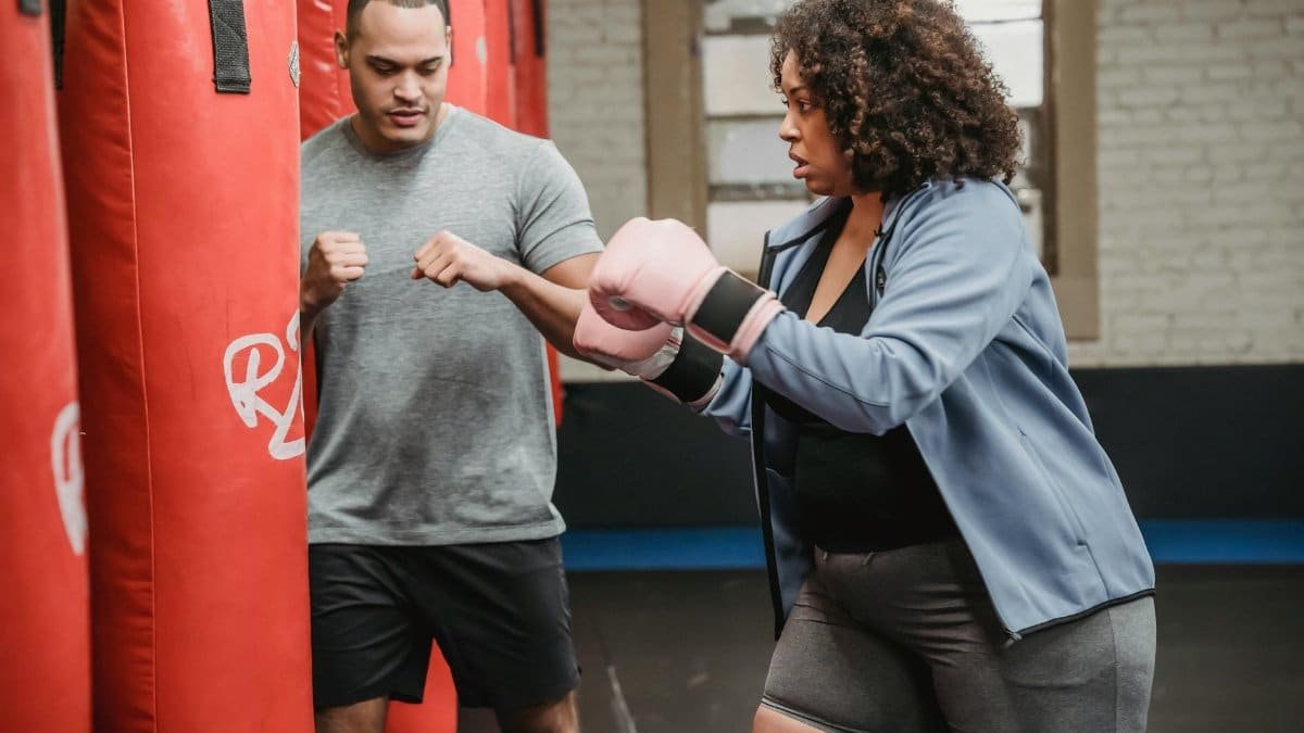 Serious ethnic coach explaining exercise to overweight African American woman punching bag in boxing gym