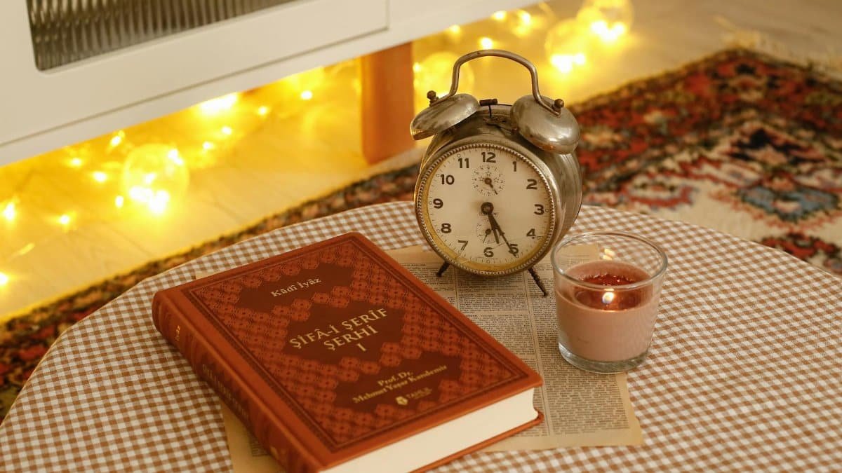 A warm, cozy still life featuring a vintage clock, book, and candle on a checkered tablecloth.