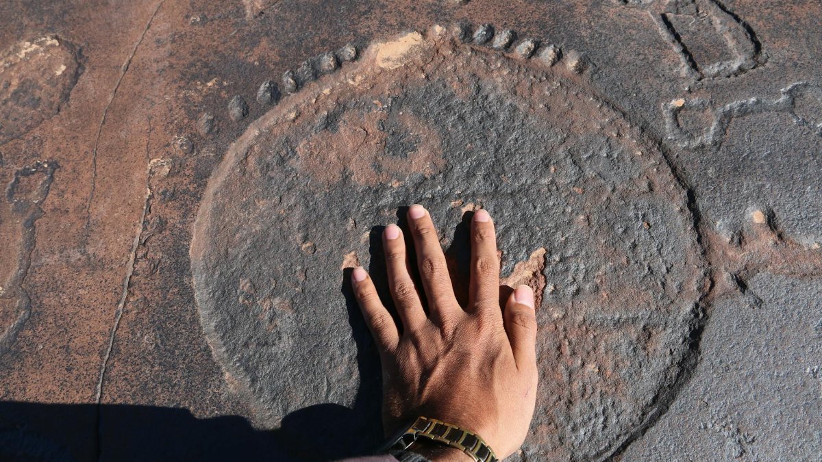 A person's hand touches prehistoric petroglyphs etched on a rocky surface outdoors.