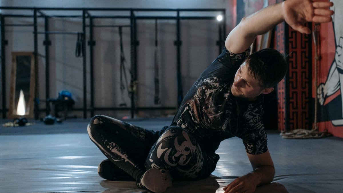 A man performs a stretching routine indoors in a mixed martial arts gym, preparing for training.