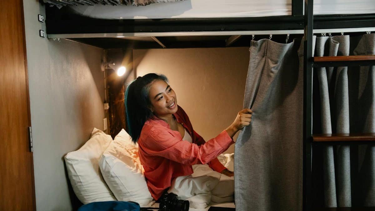 Side view of young smiling ethnic female resting in bed under white blanket with camera and phone and looking away holding curtain in train