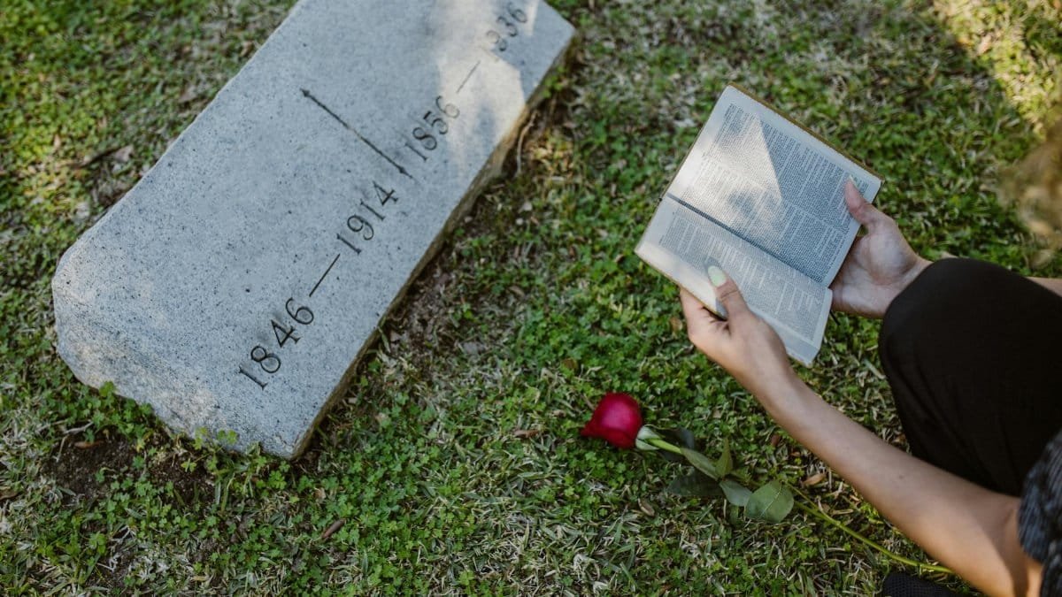 Person reading the Bible next to a gravestone with a red rose, symbolizing reflection and remembrance.