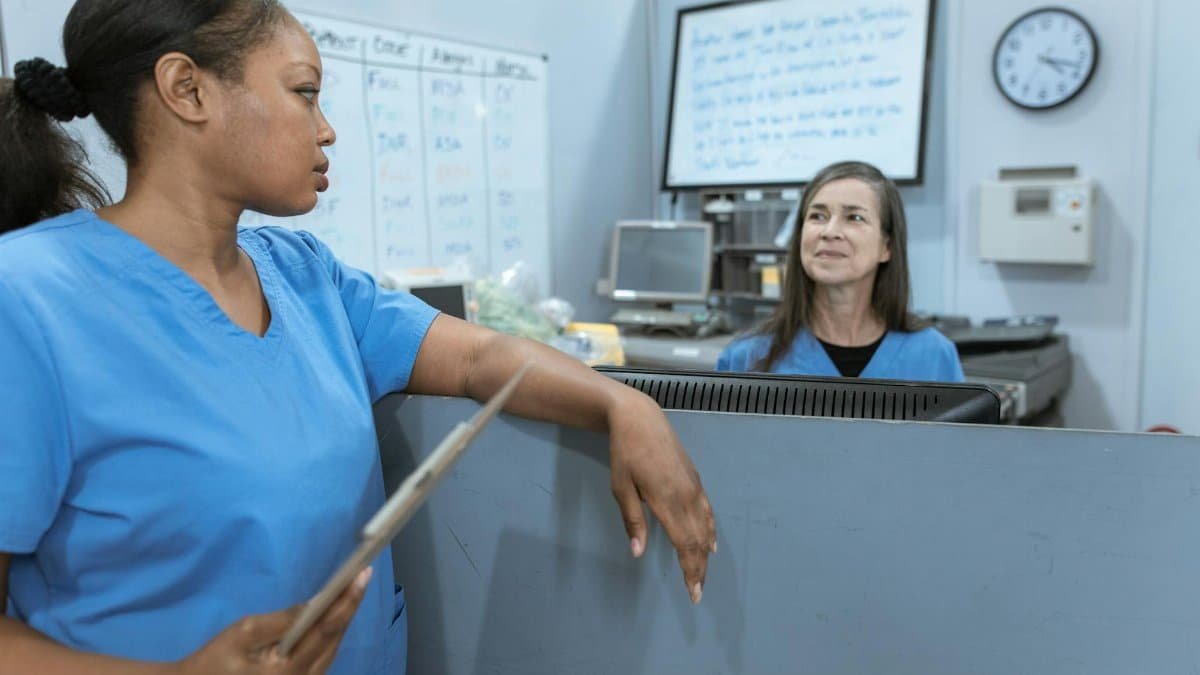 Nurses in scrub suits discussing medical tasks in a clinical environment.