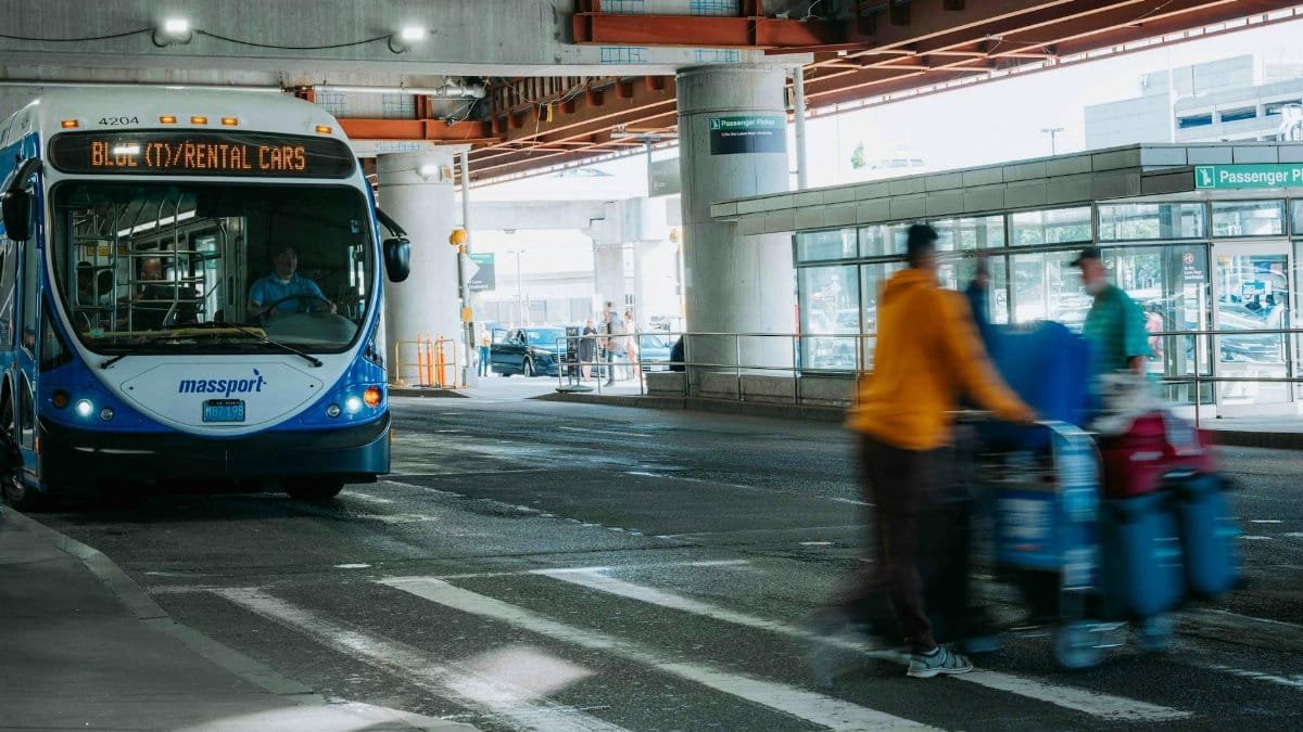 Busy airport scene showing travelers in motion and a rental car bus waiting under a covered terminal.