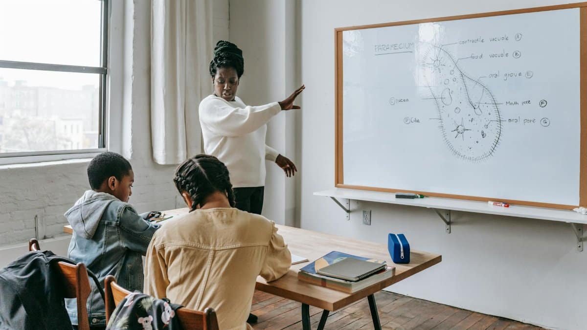 African American female teacher standing near whiteboard and giving lecture to ethnic teenagers during biology lesson