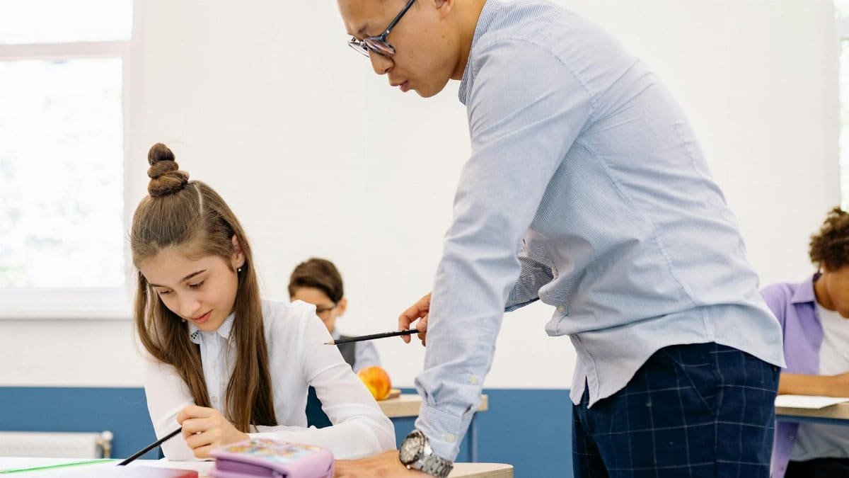 Asian male teacher assisting a young caucasian girl with her studies in a classroom setting.