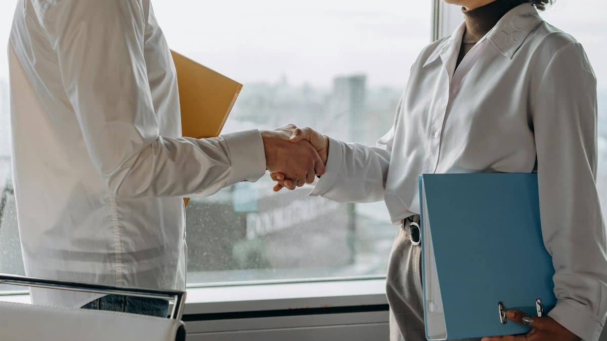 Two professionals in business attire shaking hands in an office setting with view.