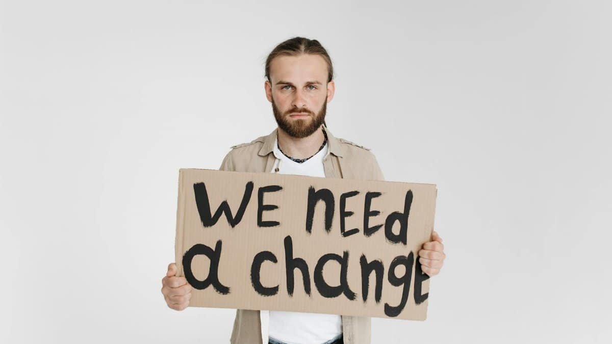 A man holding a protest sign reading 'We Need a Change' on a white background.