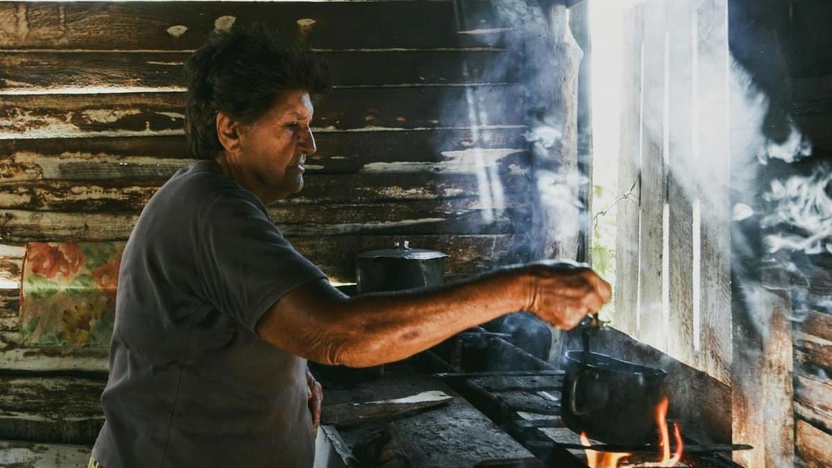 Elderly woman cooking over an open fire in a rustic wooden hut with smoke and natural light.