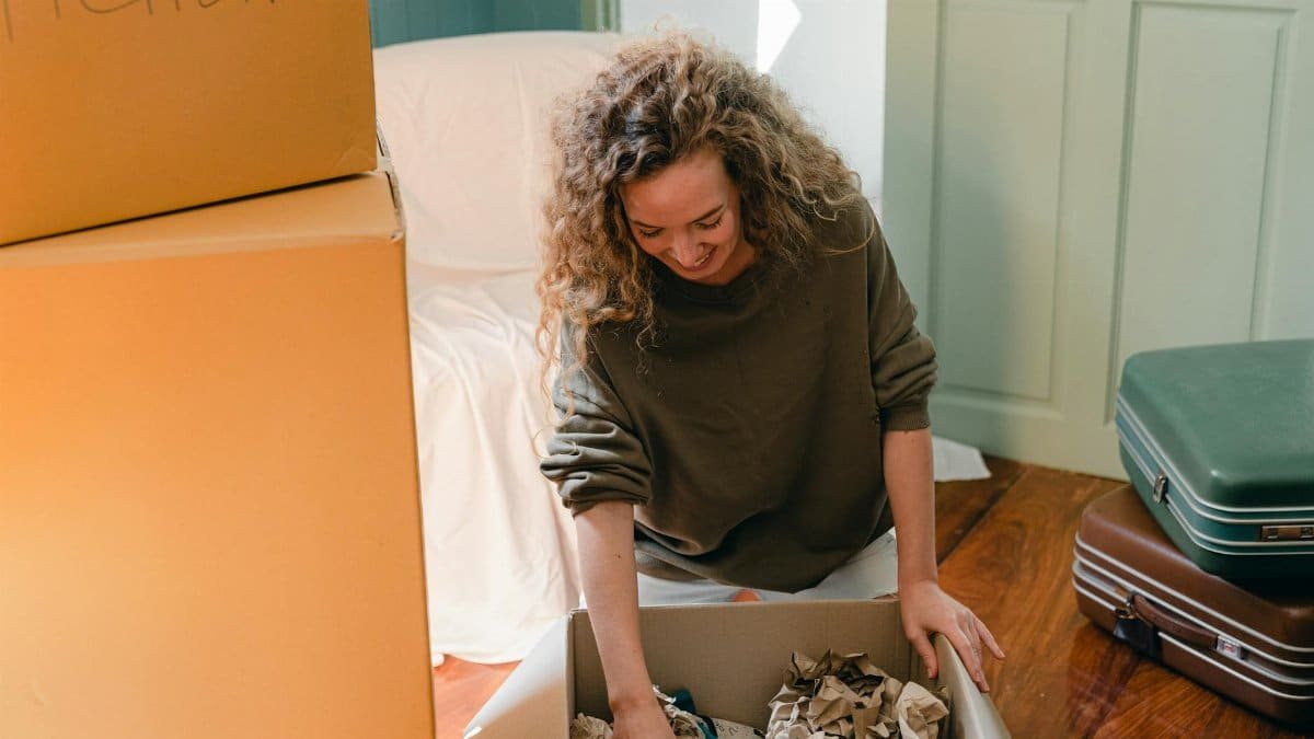 Cheerful smiling female with curly hair wearing casual sweater sitting on floor and unpacking carton box with belongings while moving into new apartment