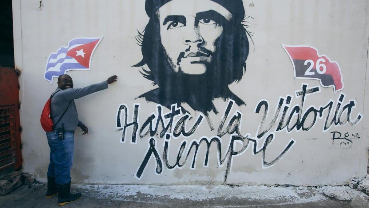 Man standing beside Che Guevara mural featuring Cuban flag and symbol of the 26th of July Movement.