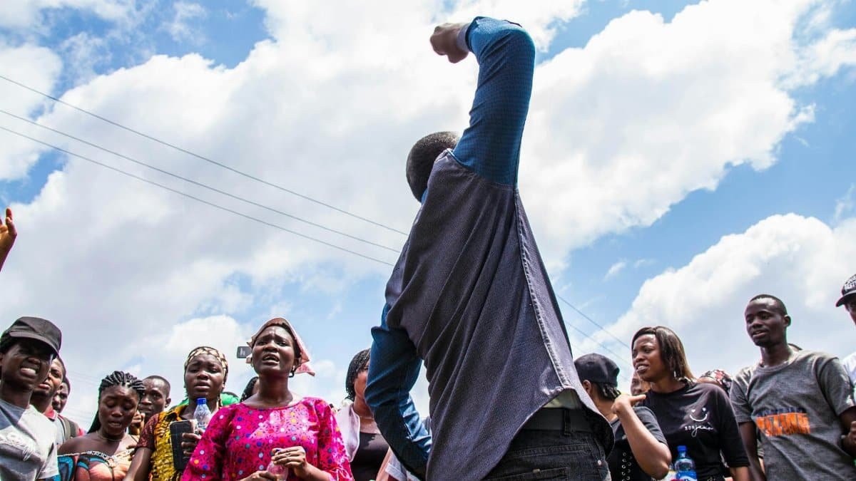 A diverse group of people engaged in an outdoor protest with raised voices and arms, displaying unity and motivation.