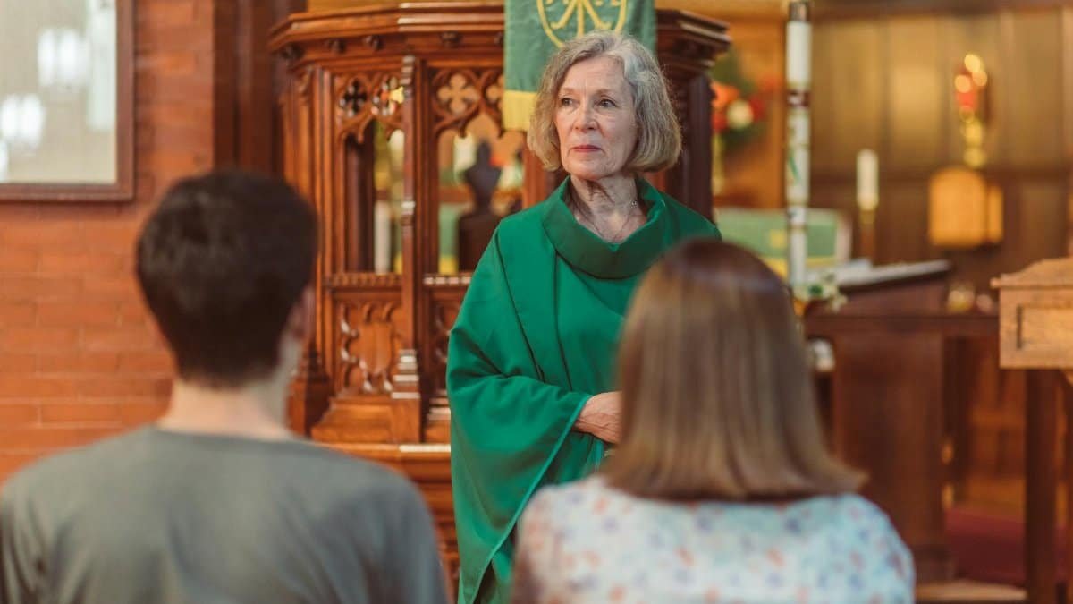 Elderly priest in green cassock leads a church service, engaging parishioners in an elegant wooden interior.
