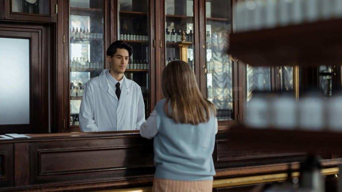 A pharmacist assists a customer at a vintage wooden pharmacy counter indoors.