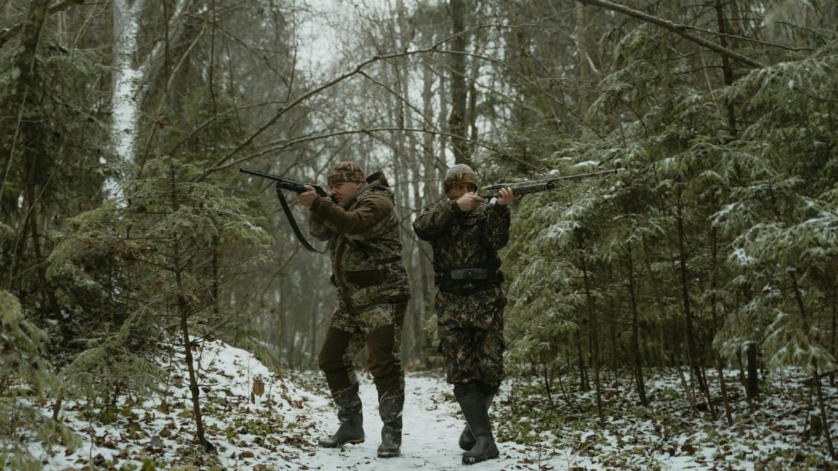 Two hunters in camouflage walking through a snowy forest with rifles.