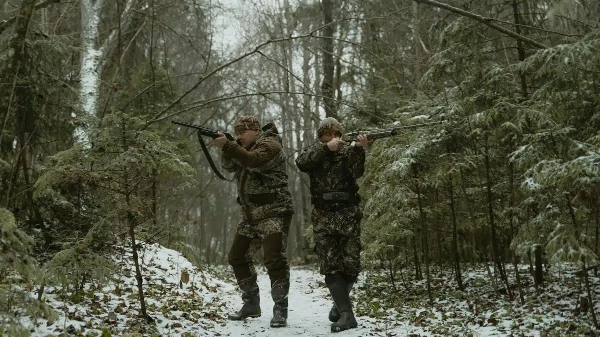 Two hunters in camouflage walking through a snowy forest with rifles.