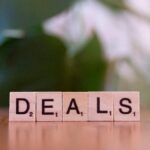 Close-up of Scrabble tiles spelling 'DEALS' on a wooden surface with blurry green background.