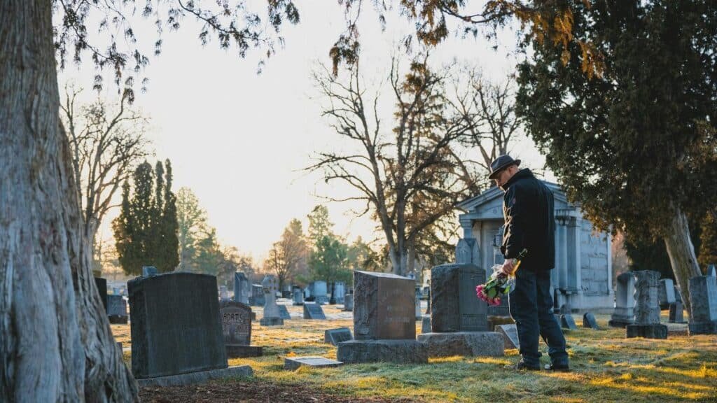 Elderly man honors a loved one with flowers in a serene cemetery setting during daylight.