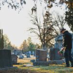 Elderly man honors a loved one with flowers in a serene cemetery setting during daylight.