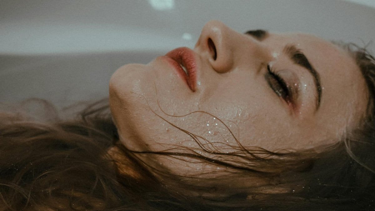 Relaxed female with closed eyes and smudges makeup on face lying in bathtub with hair in water at home inside