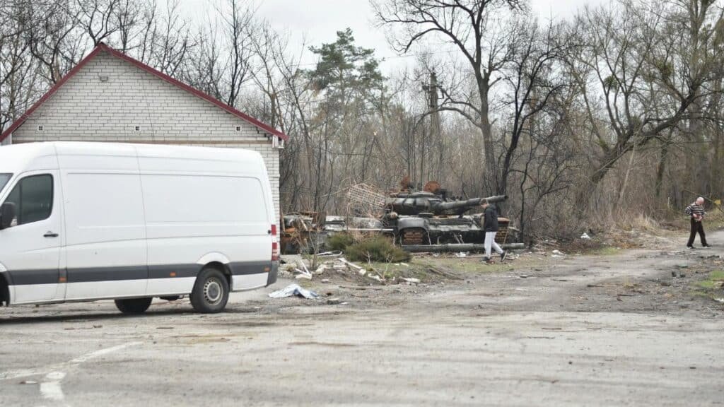Scene of a war-torn area with an abandoned tank and van, illustrating the aftermath of conflict.