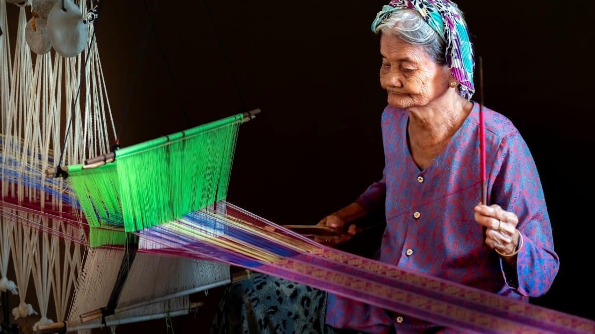 Elderly woman skillfully weaving colorful textile on a loom inside a dim room.