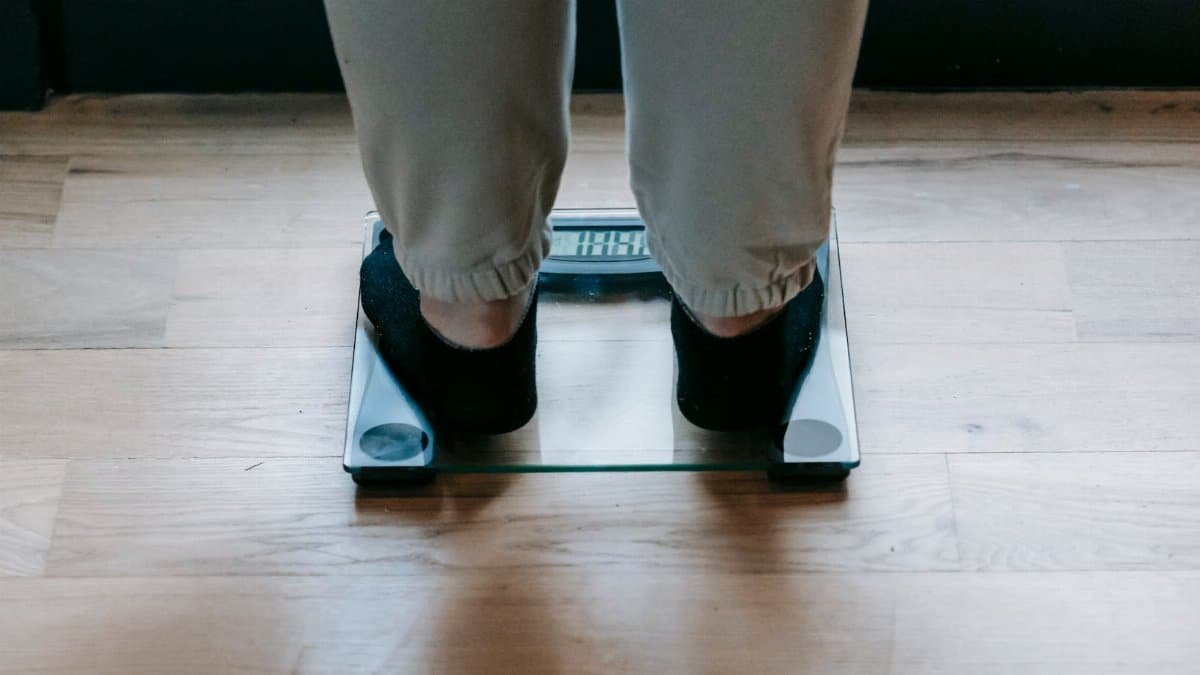 Back view of crop faceless obese female in socks weighing on glass scale placed on wooden floor in light room in daytime