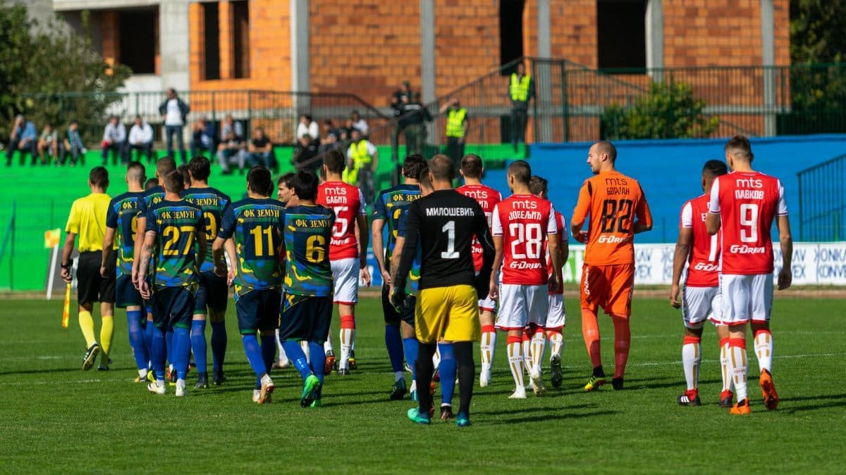 Players from two teams in blue and red uniforms preparing for a football match on a green field.