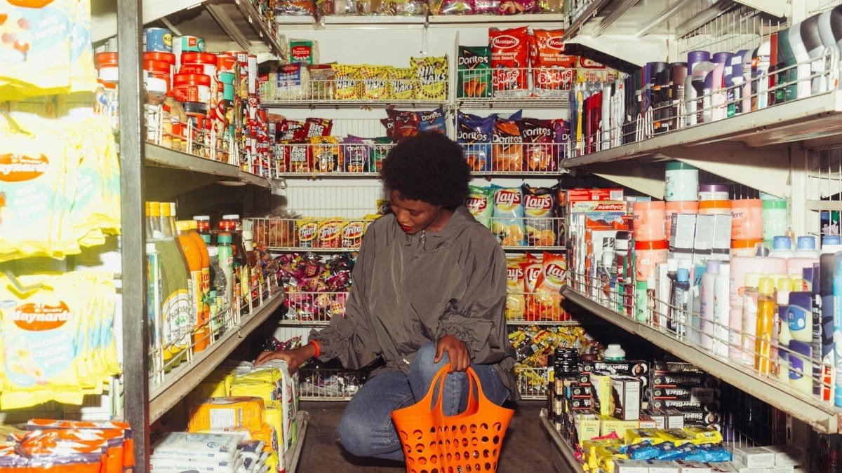 Full length of adult ethnic female buyer selecting goods while squatting with shopping bag among grocery shelves