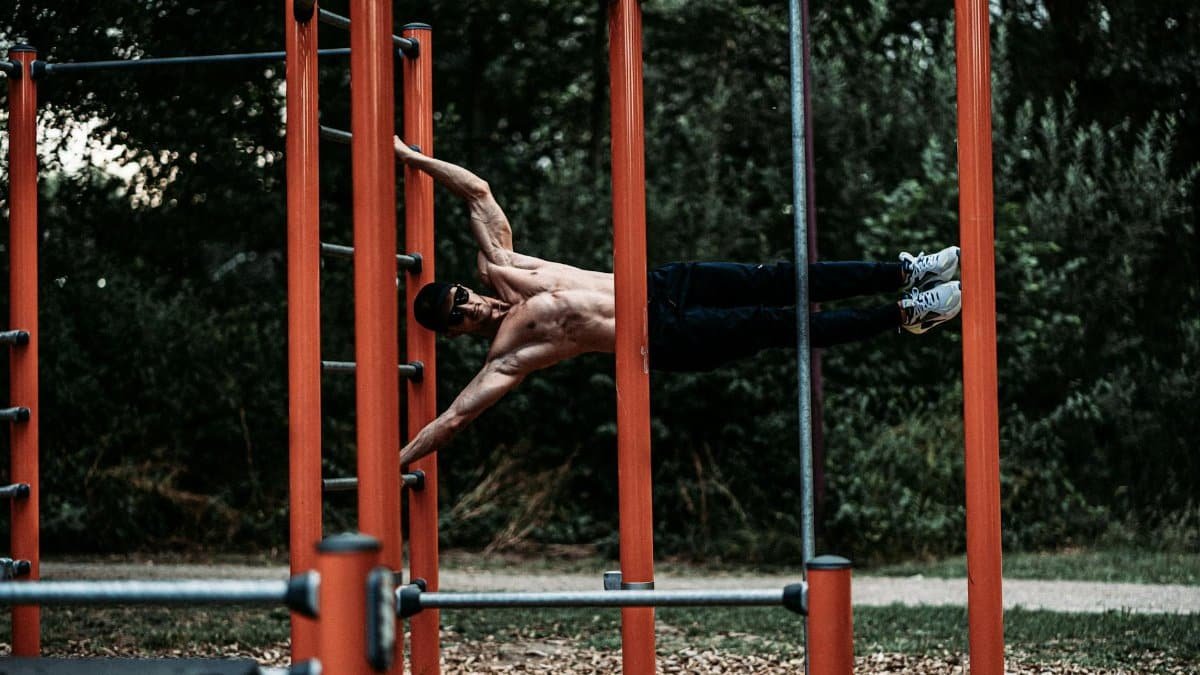 A shirtless man demonstrates strength and agility on outdoor calisthenics bars.