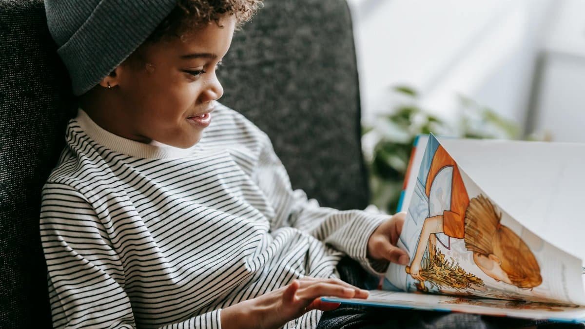 Side view of crop glad African American boy with curly hair smiling and watching pictures of book on blurred background