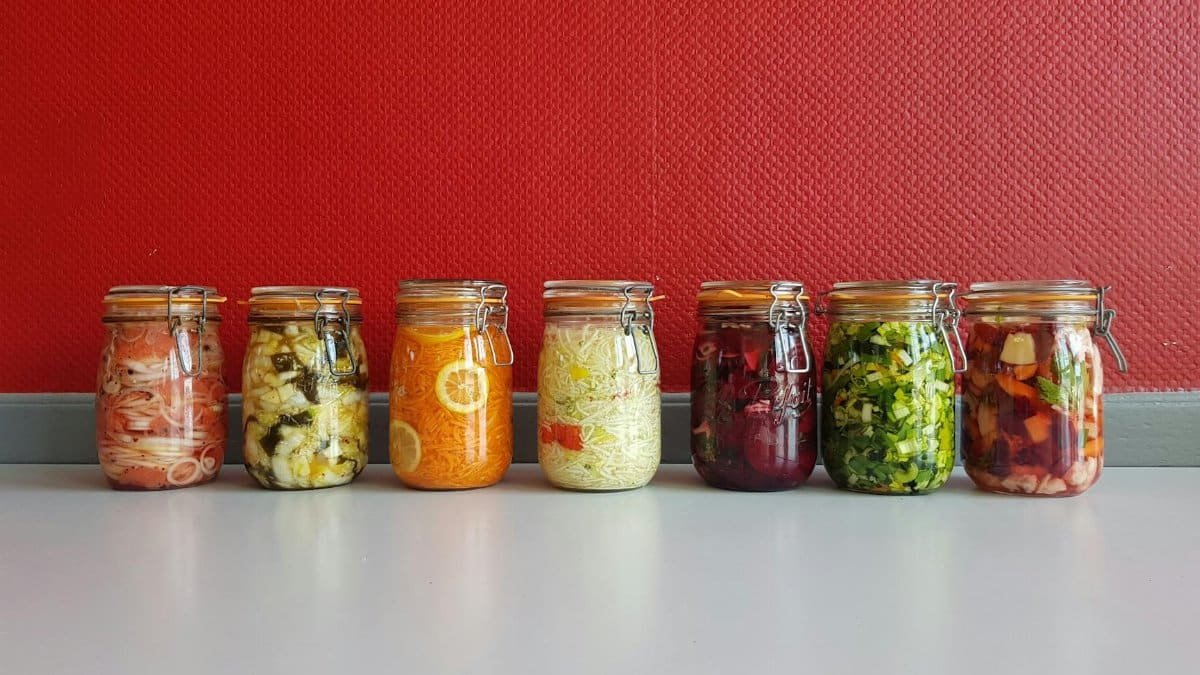 A row of vibrant, multicolored mason jars filled with assorted fermented vegetables against a red wall.