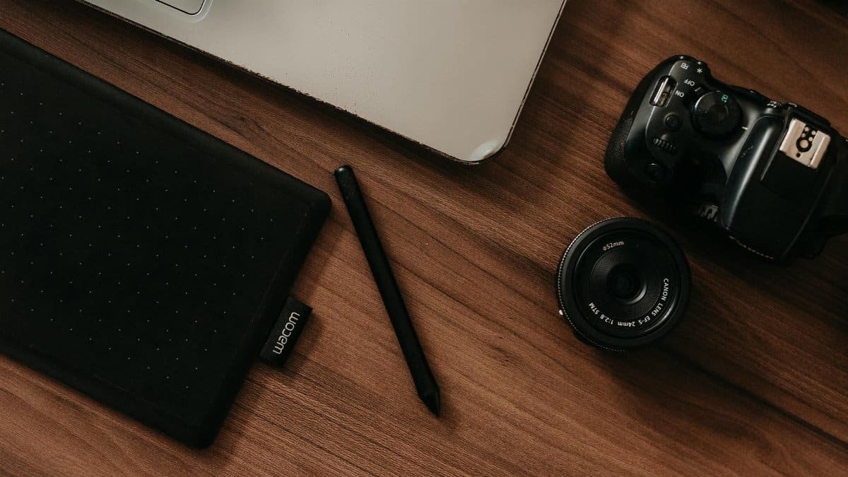 A top view of a digital workspace featuring a camera, tablet, pen, and laptop on a wooden desk.