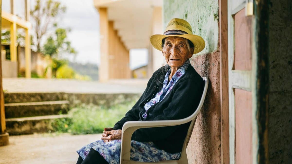 Senior woman in a hat sitting outdoors on a chair, showcasing peaceful rural life.