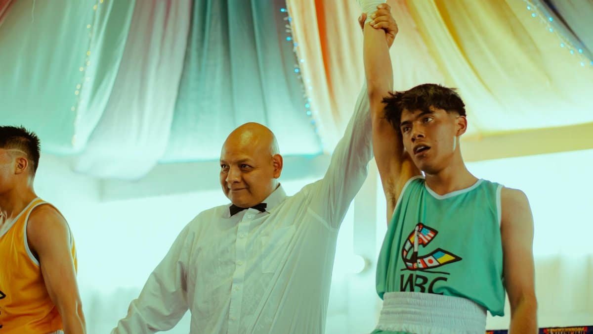 A triumphant teenage boxer winning a match, his hand raised by a referee, in a vibrant indoor setting.