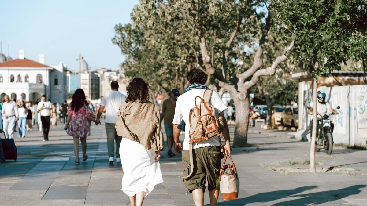 A couple strolls down a bustling city street surrounded by lush trees and vibrant urban life.