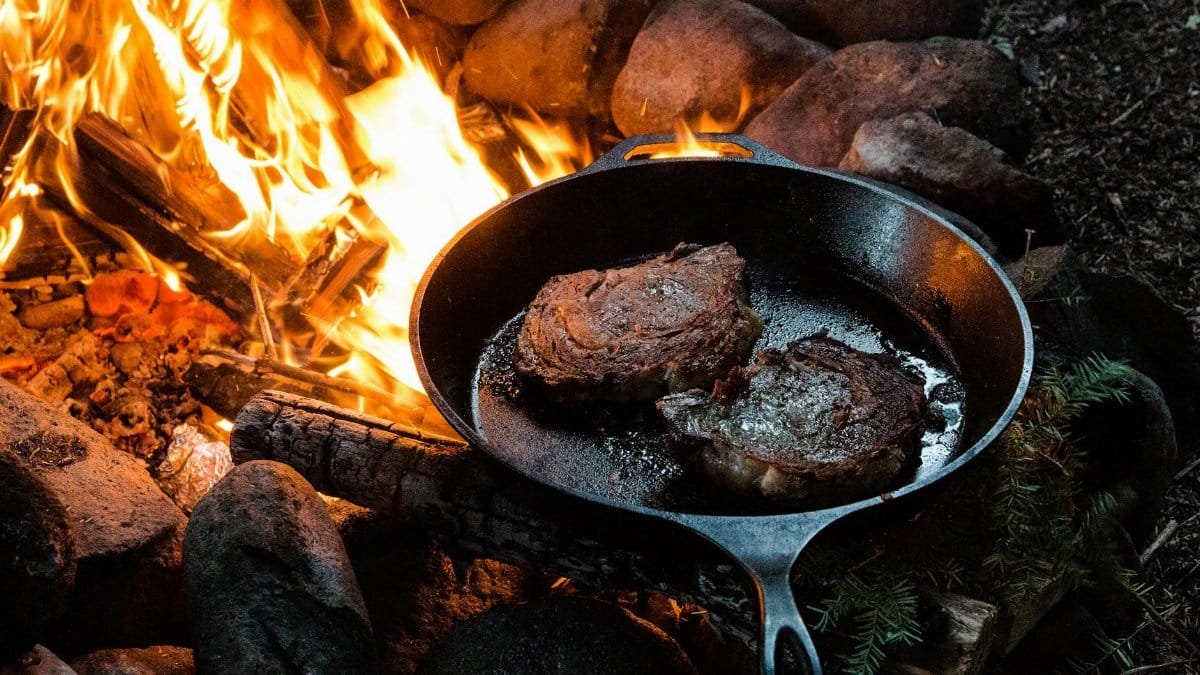 Sizzling steaks cooking in a cast iron pan over a campfire surrounded by logs and rocks.