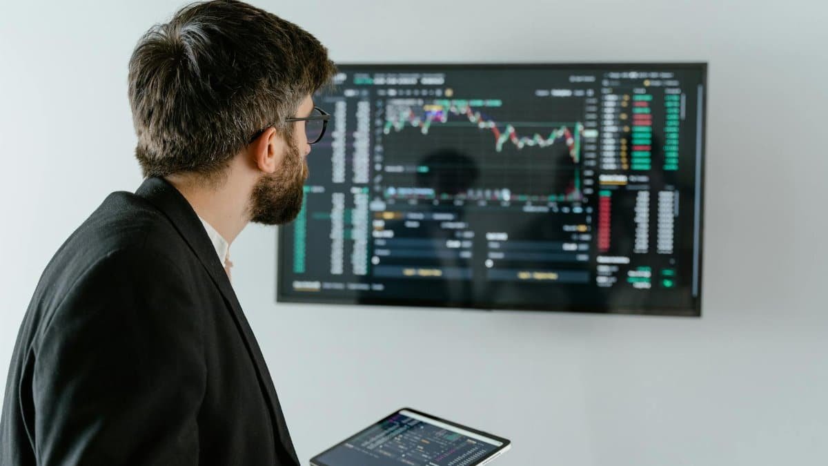 A businessman examines stock market data displayed on a monitor, holding a tablet.