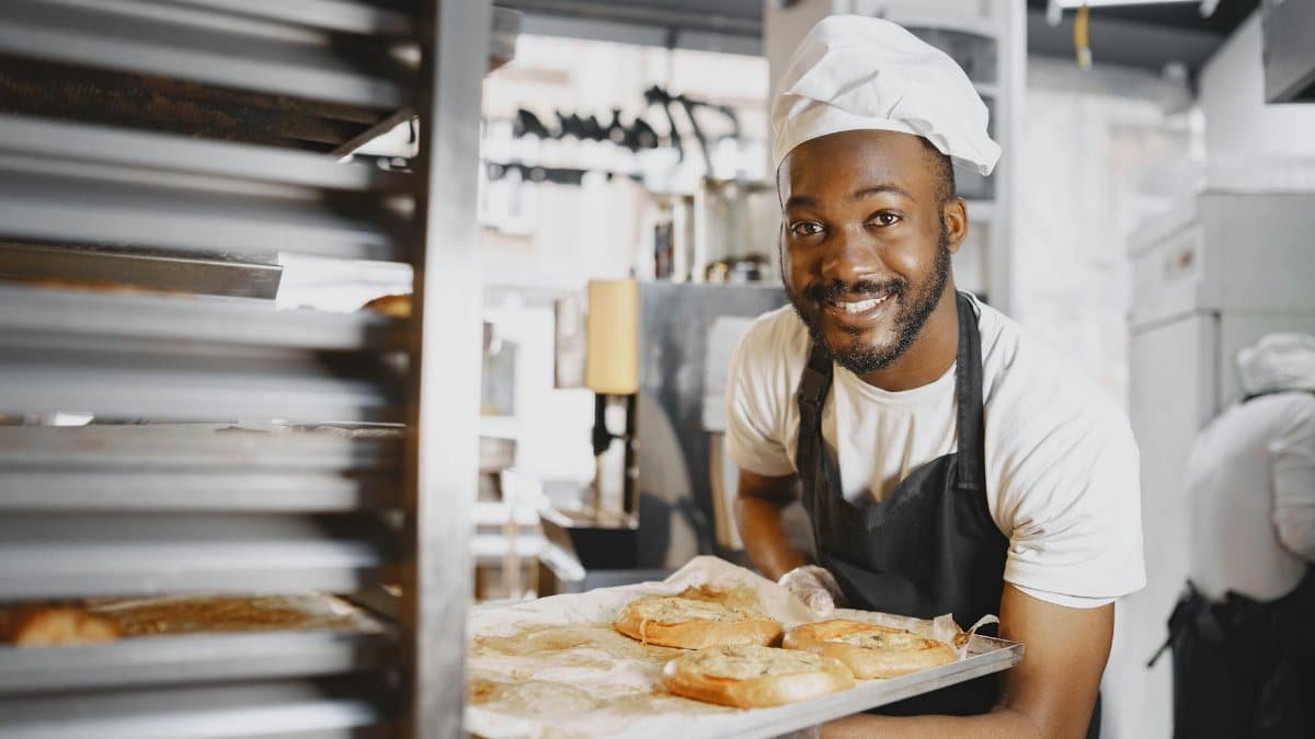 A cheerful baker presenting fresh breads from an industrial oven. Captured in a bakery setting.