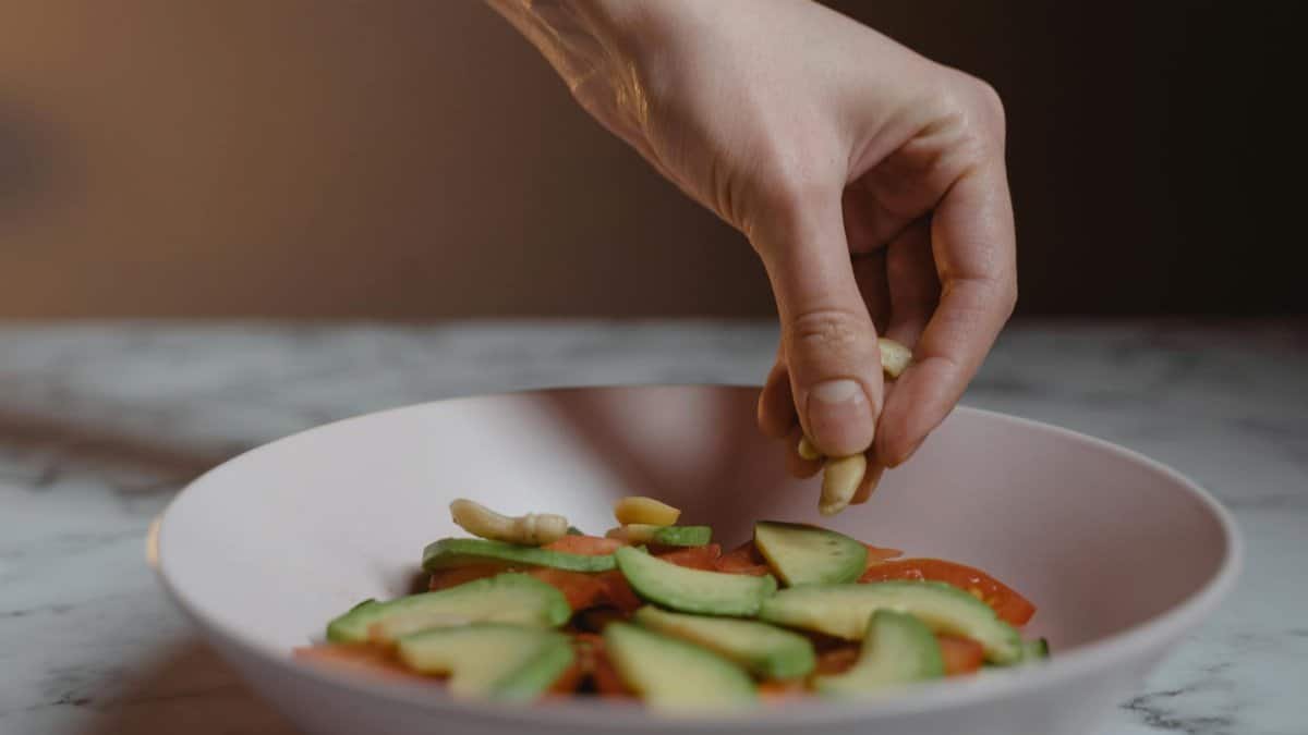 Hand adding cashews to a fresh vegan salad with avocado slices in a ceramic bowl.