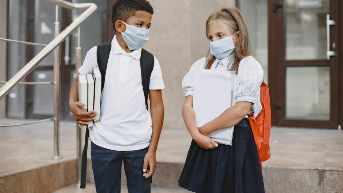 Two school children wearing face masks, holding books, standing outside a school building.