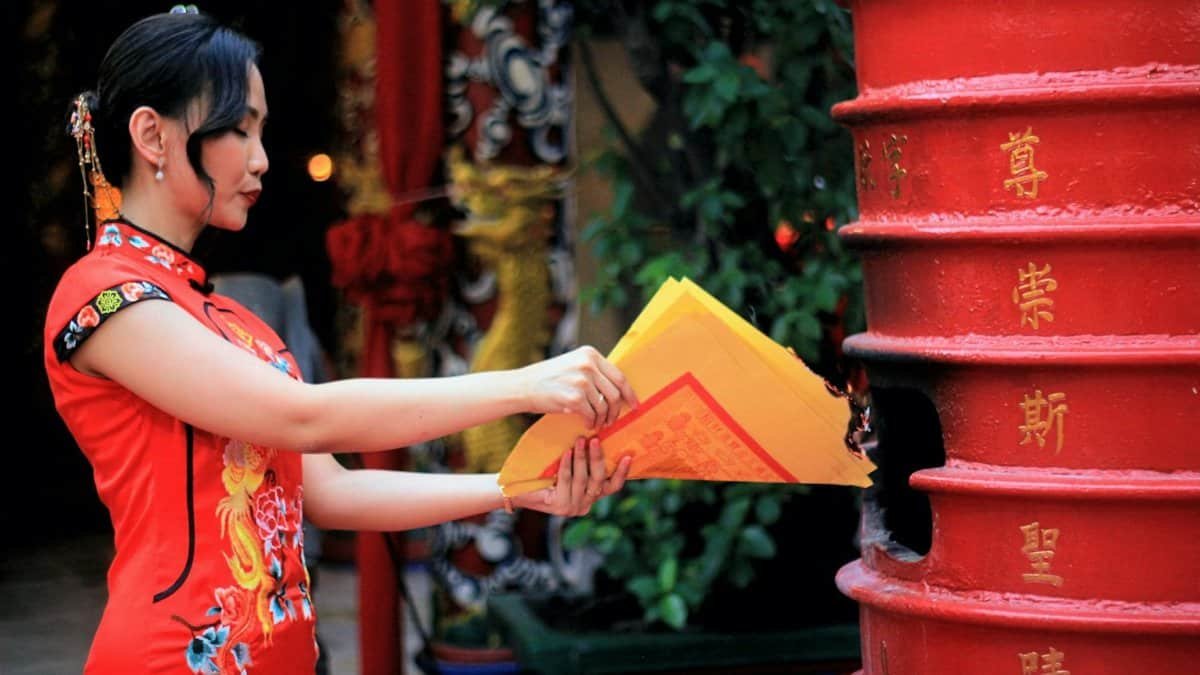 Asian woman in red qipao ceremonially burns joss paper during a traditional event.