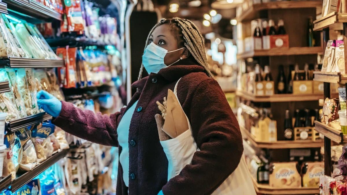 Side view of African American female shopper in protective mask and gloves selecting goods in grocery store
