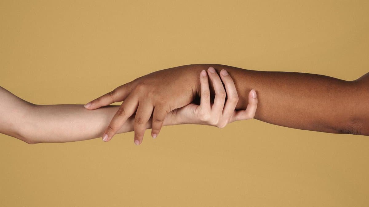 Crop anonymous multiracial females showing support and respect by holding hands tightly against yellow background