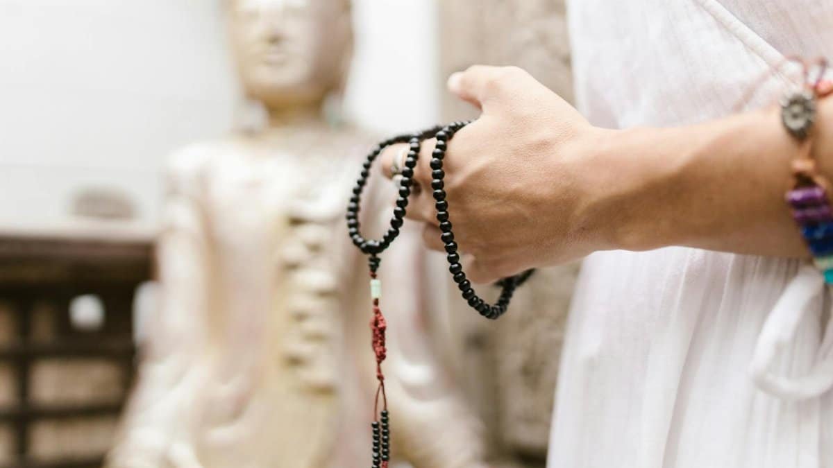 A close-up of a hand holding black prayer beads near a blurred Buddha statue, symbolizing spirituality.